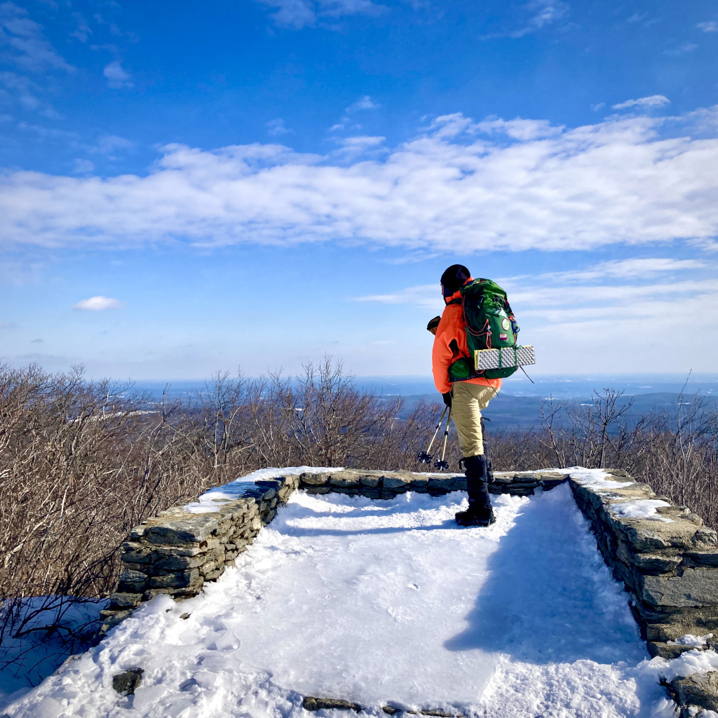 Hiker standing on a snow covered observation overlook with blue skies and mountain ranges in far distance