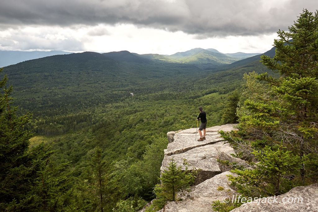 a solo hiker looks out over a forested valley towards a range of mountains in the distance. He is standing on a outcropping of ledge that drops off to the valley floor