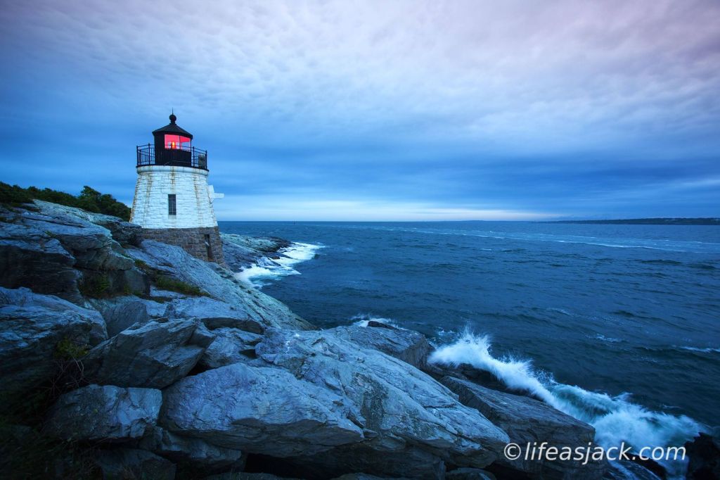 A white granite lighthouse nestled in shore ledge by a blue sea. The red warning beacon can be seen in the lighthouse tower. waves crash on the rocks below the lighthouse with white spray.