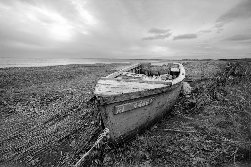 A wooden skiff is ashore on a beach of straw, seagrass and shells. The sea is in the background. The image is black and white.