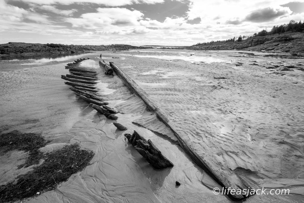 The weathered spine and beams of a wooden sailing ship poke up from the sand on a rocky beach.