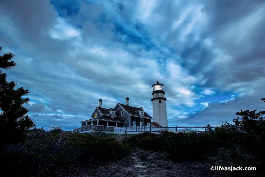 A deep blue cloudy sky at nighfall. Below a lightkeepers house with a white lighthouse tower flashes a white beacon from it's lens. A window in the keepers house glows with a warm yellow colr.