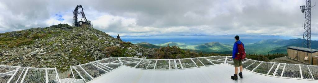 A hiker standing on a helicopter pad on top of a mountain. his back is to the camera. he is looking at a cell phone communications tower that has been destroyed and is bent and collapsed.