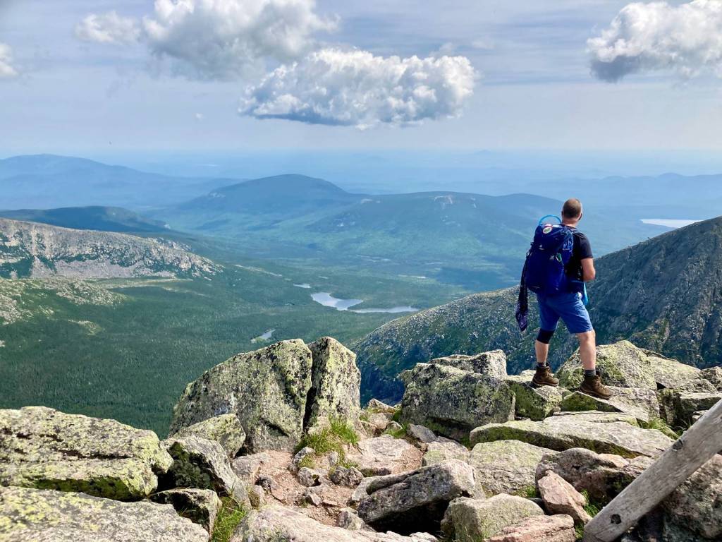 A hker on the summit of a mountain. He is in the right corner of the image, with his back to the camera looking out to the horizon on a sunny day with blue skies and clouds in the distance.