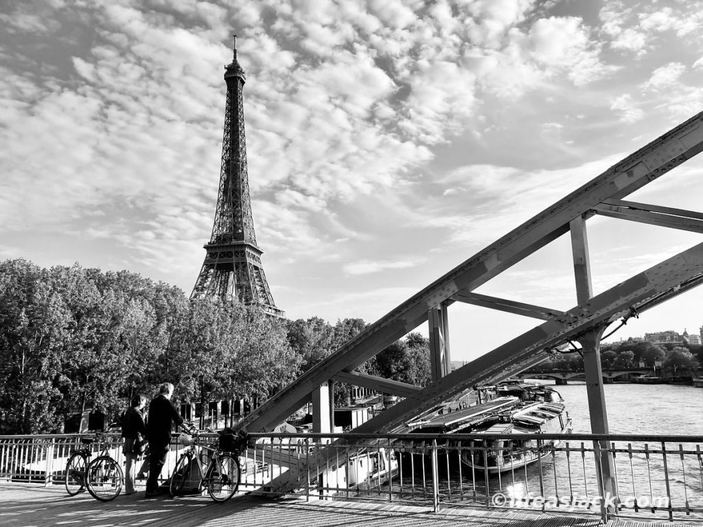Two cyclists stand on a bridge overlooking the Seine river in Paris, France. The Eiffel Tower can be seen in the background.