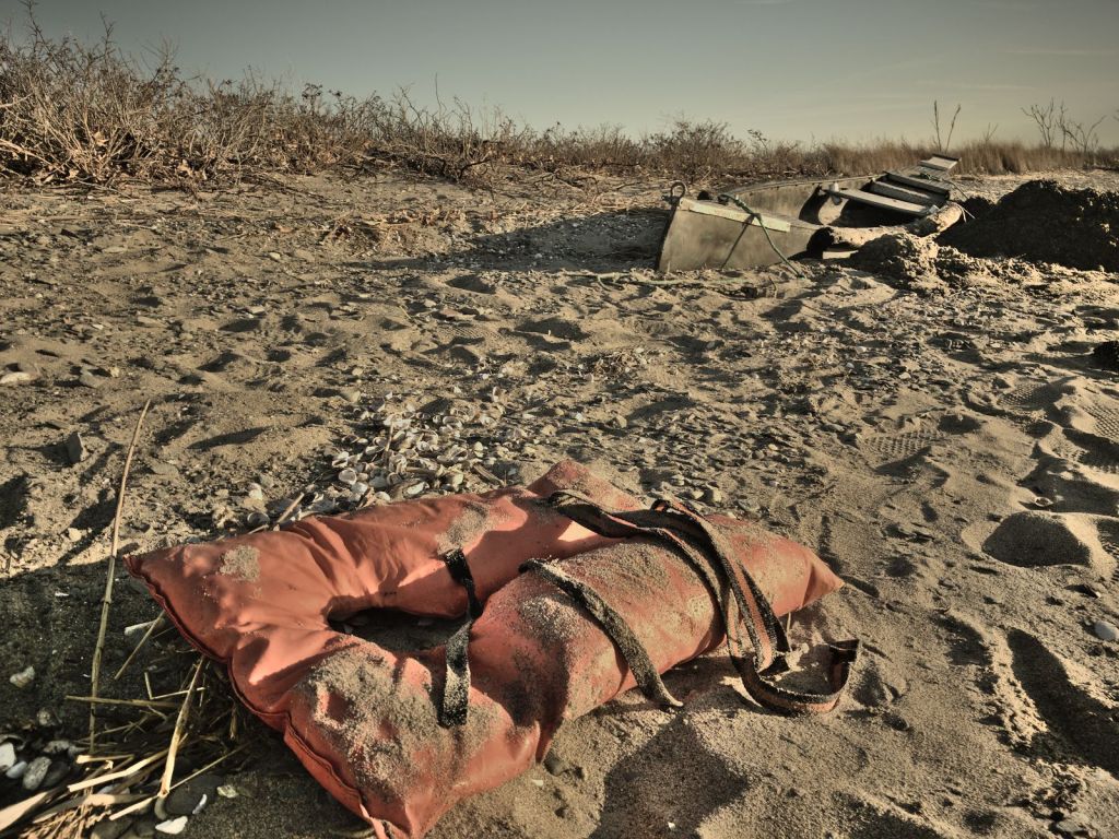 A derelict kayak is half buried in beach sand, abandoned. In the foreground is a tattered life vest, crusted with sand.