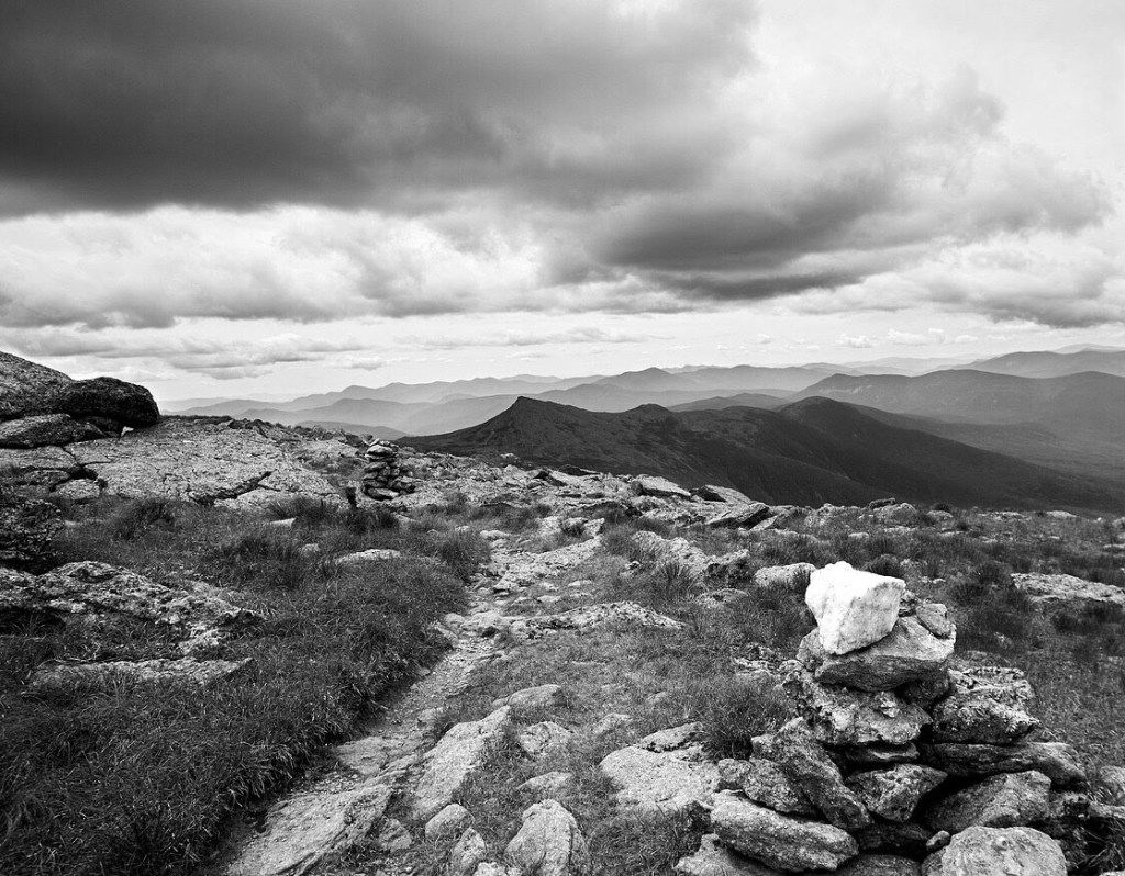 A black and white image of a hiking trail in the white mountains of New Hampshire. The foreground is studded with heavy granite boulders. A dark mountain range is seen in the distance under dark storm clouds.