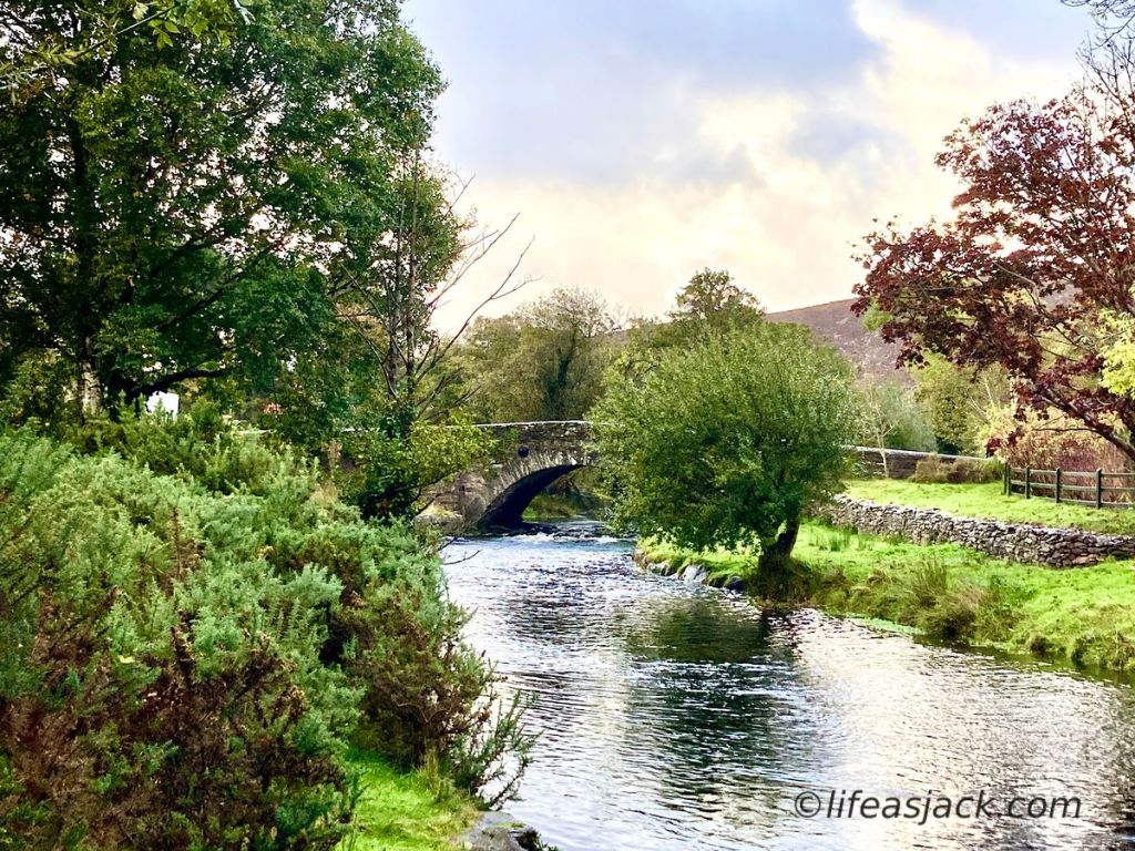 A stone bridge centered in the image crosses a stream, framed in green grass and leafy green trees.