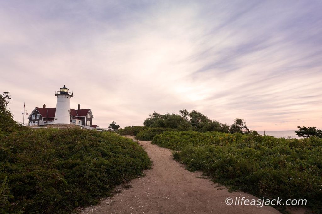 a sandy path in the foreground center of the image is bordered by green ground cover. The path leads to a white lighthouse and keepers house with a red roof. The sky is overcast at dawn with a faint glow of the rising sun.