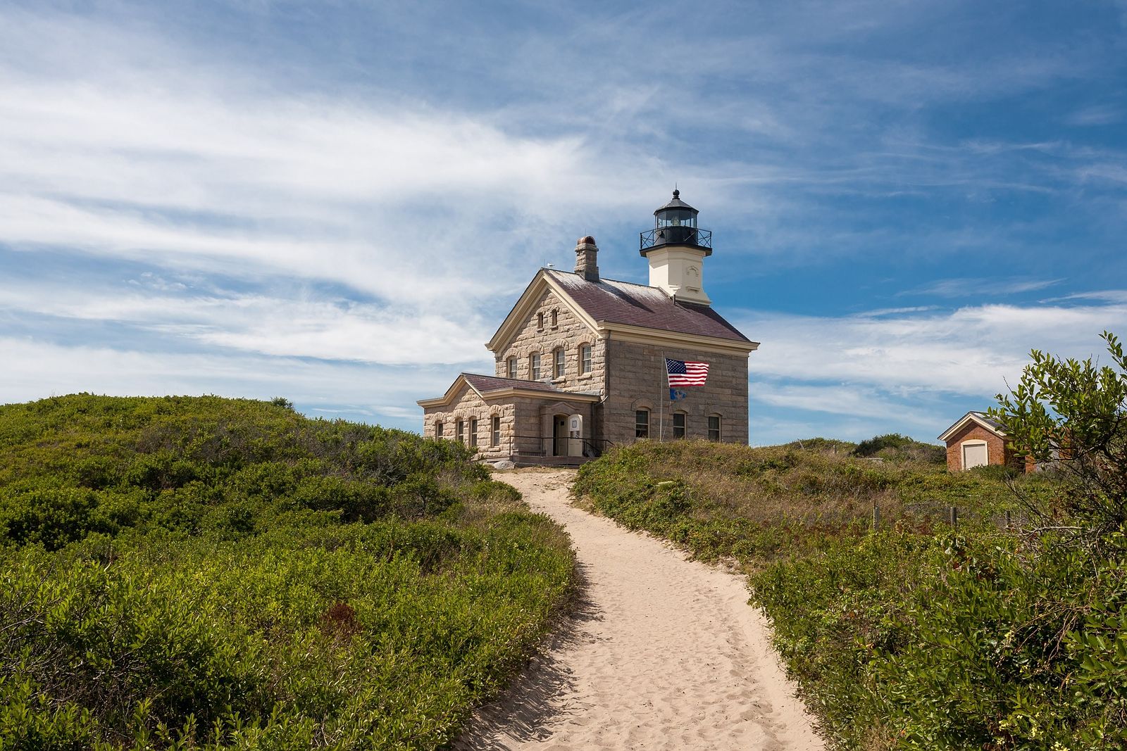 A granite lighthouse is centered on the frame under a blue sky. a sand path leads to the lighthouse surrounded by green sea grass and shrubbery.