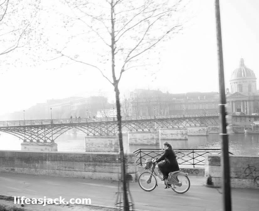 A black and white image of a woman riding a bicycle along the River Seine, Paris France on a foggy morning.