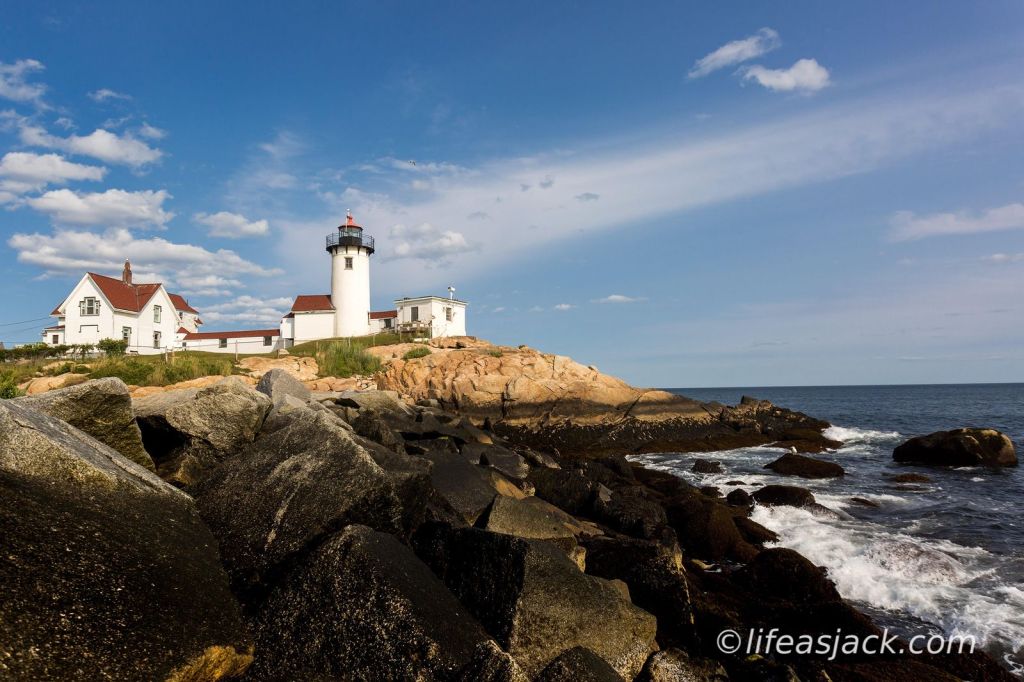A white lighthouse and keepers house with a red roof sits in the left corner of the image under a blue sky. rocky shoreline in Gloucester Harbor is in the foreground. Ocean waves break on the rocks from the right side of the image.