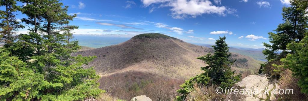 a mountain covered in brown trees is center in the frame. green fir trees frame the mountain on both sides of the picture. A blue sky with a few scattered clouds are over the peak.