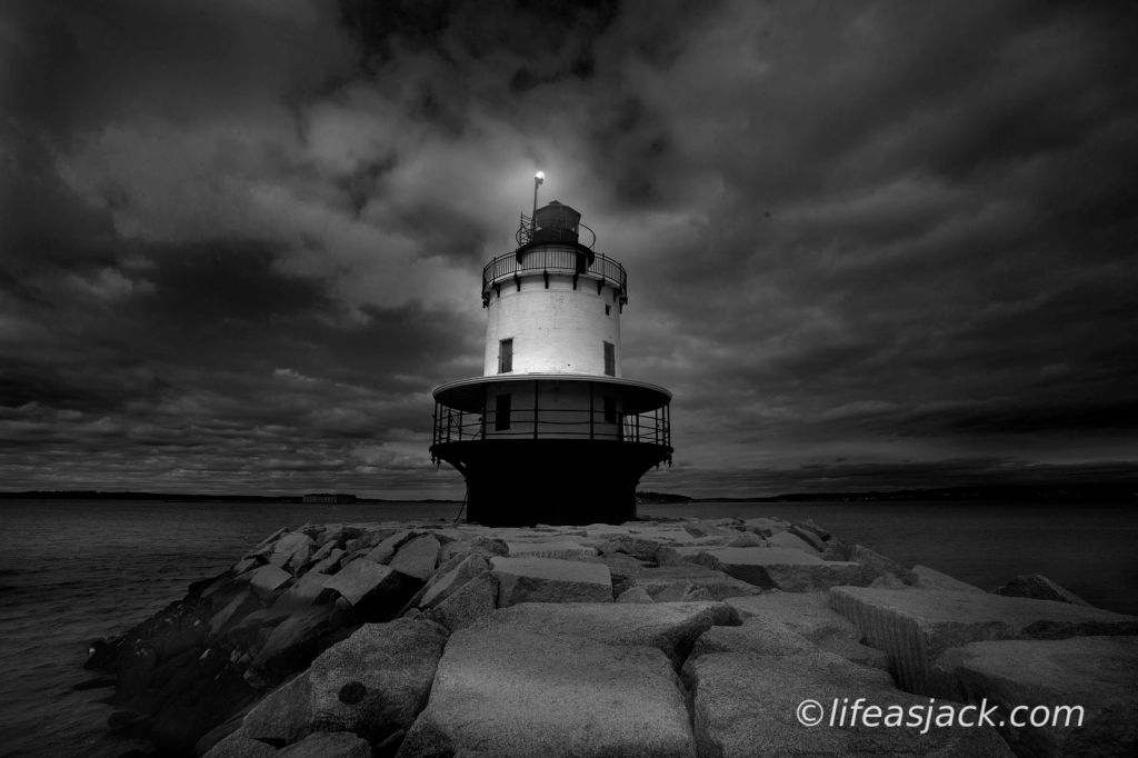 A lighthouse centered in the image on a jetty of granite boulders. The lighthouses beacon is illuminated above the tower. The sky is overcast at night with dark clouds.