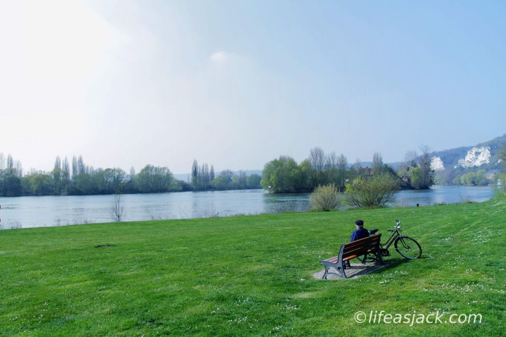 a man sits on a park bench overlooking a river. his bicycle is parked near him, resting on the bench.
