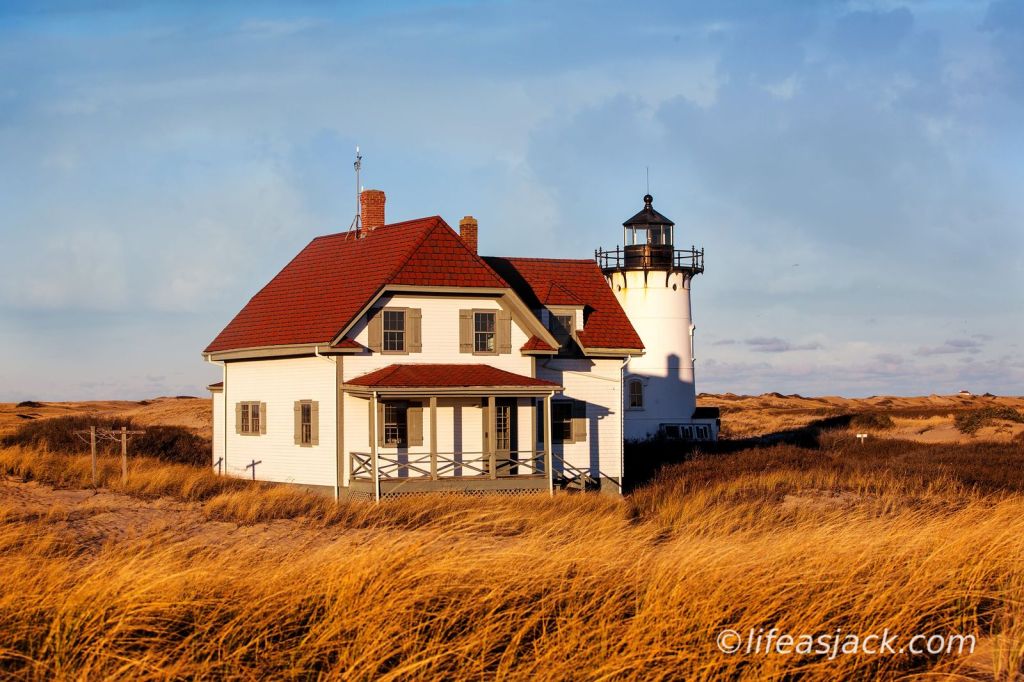 A white lighthouse with a black turret is attached to a white keeper's house with a red roof. The house is surrounded by tall yellow sea grass blowing in the wind under a blue sky.