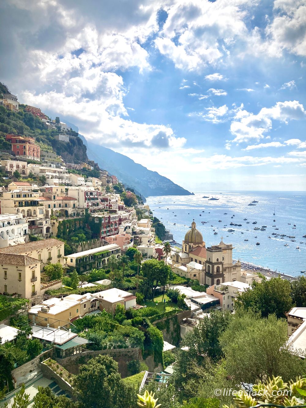 houses perch on steep cliffs in the left of the image frame. A blue sky shines on a crowded harbor in the right side of the image.
